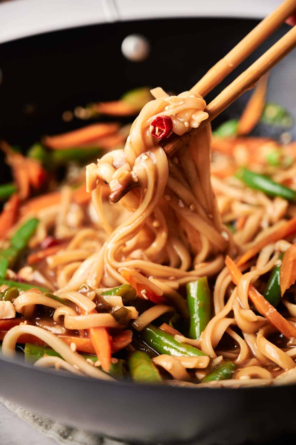 Chopsticks lifting spicy noodles with vegetables from a pan, featuring sliced carrots, green beans, and savory sauce.
