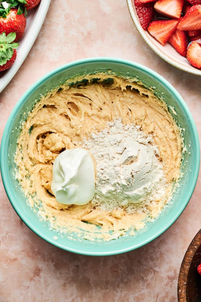 A green bowl containing cookie dough with a dollop of cream and flour on top, evoking the beginnings of a delightful strawberry cake. Surrounding it are dishes of fresh strawberries on a marble countertop.