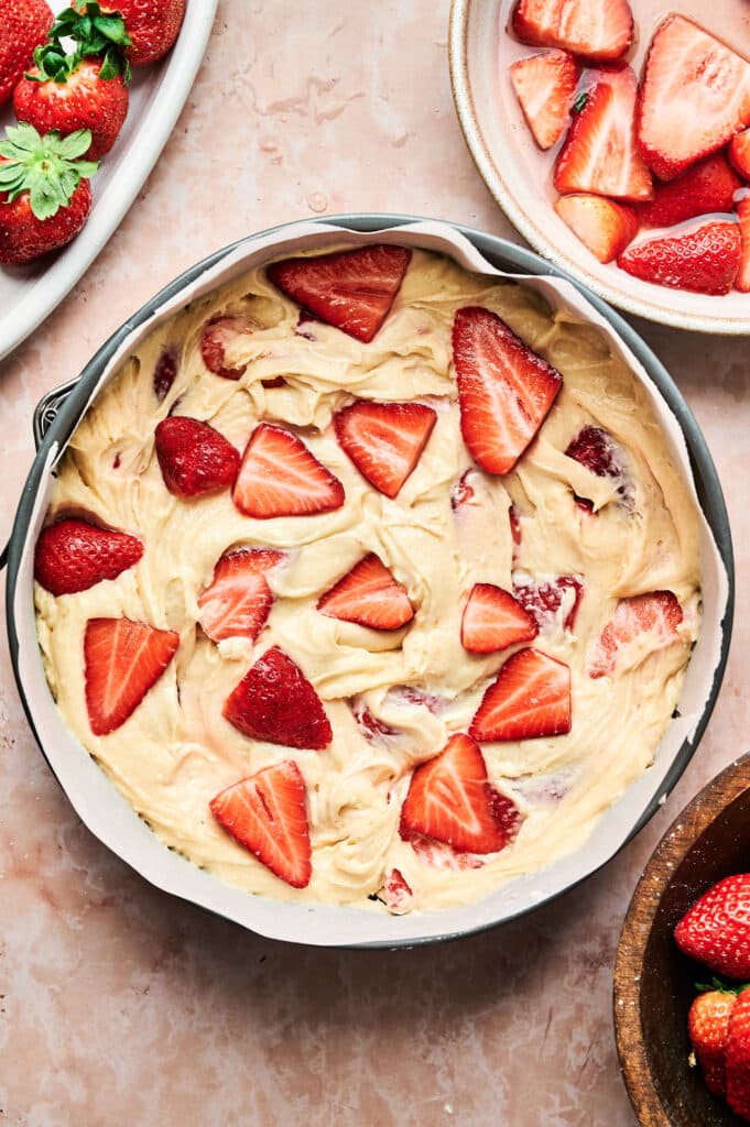A strawberry cake in the making: a round pan filled with cake batter, topped with sliced strawberries, surrounded by bowls of fresh berries on a marble surface.