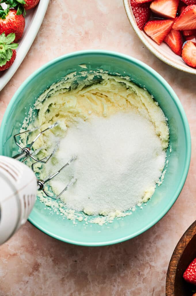 An electric mixer in a green bowl with sugar and butter on a pink countertop is surrounded by sliced strawberries, hinting at the creation of a delectable strawberry cake, alongside a plate of whole strawberries ready to be transformed.