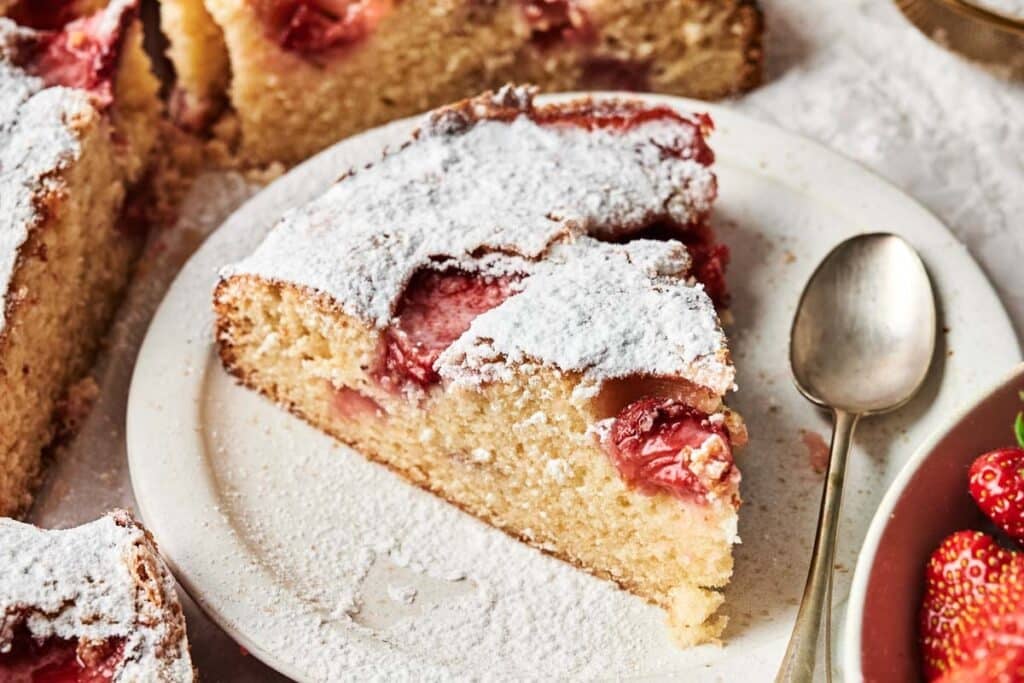 A slice of strawberry cake dusted with powdered sugar sits on a white plate next to a spoon and a scoop of creamy Biscoff Ice Cream.