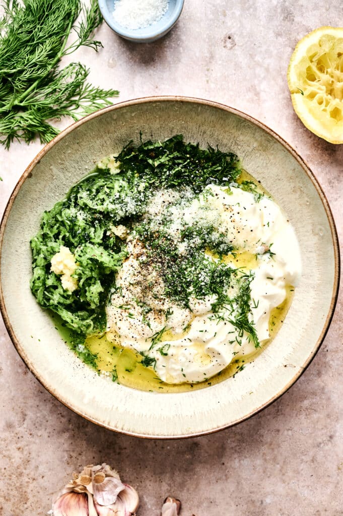 A bowl of tzatziki with yogurt, grated cucumber, chopped dill, minced garlic, olive oil, pepper, and salt on a stone surface, surrounded by fresh dill, lemon, garlic, and a small dish of salt.