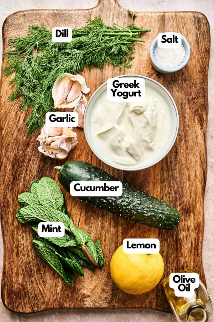 A wooden board displays dill, garlic, mint, cucumber, lemon, Greek yogurt, salt, and a small bottle of olive oil&mdash;each labeled as classic ingredients for making homemade Tzatziki.