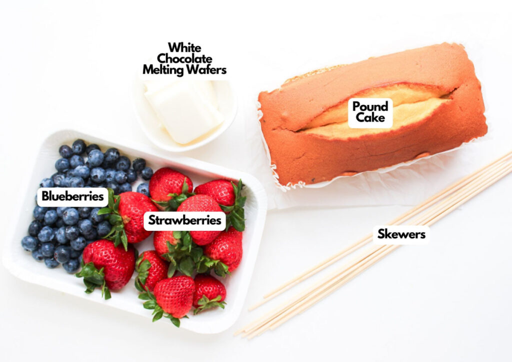 Tray with blueberries, strawberries, white chocolate melting wafers, a loaf of pound cake, and several wooden skewers&mdash;everything you need to make delicious 4th of July Fruit Kabobs&mdash;arranged on a white surface.