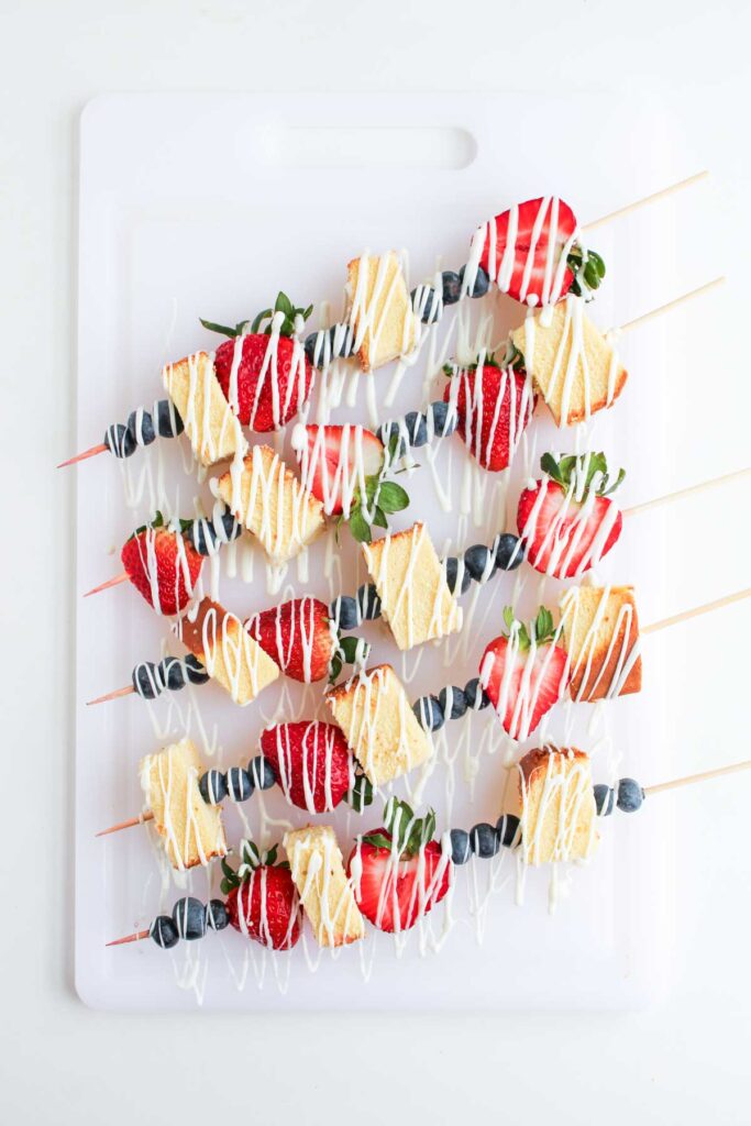 4th of July Fruit Kabobs featuring skewers with strawberries, blueberries, and pound cake cubes, drizzled with white icing, are arranged on a white cutting board.