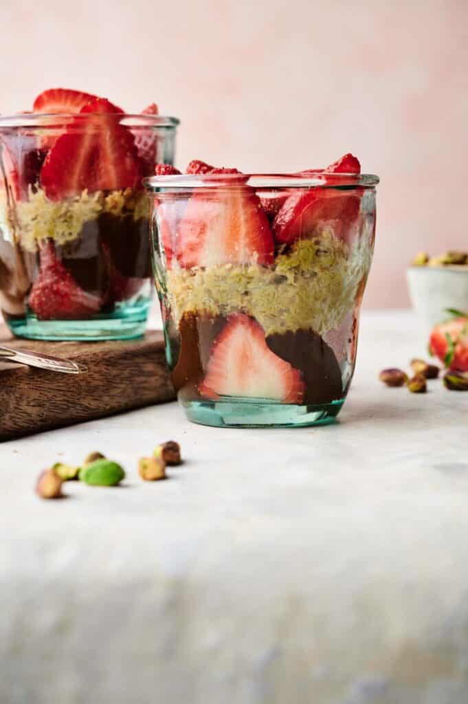 A glass jar filled with layers of chocolate, oats or chia pudding, and sliced strawberries creates a tempting Dubai Chocolate Strawberry Cup, with another similar jar in the background. Pistachios and a strawberry rest on the table.