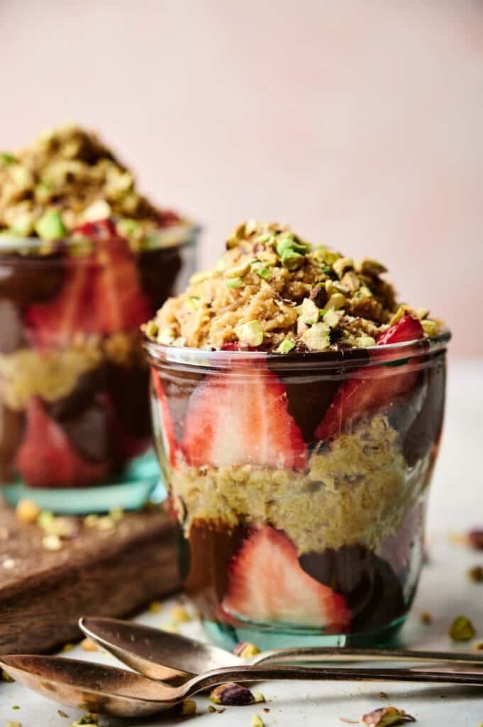 Dubai Chocolate Strawberry Cups: Glass cups layered with sliced strawberries, chocolate pudding, quinoa, and chopped pistachios, with two spoons placed nearby on a white surface.