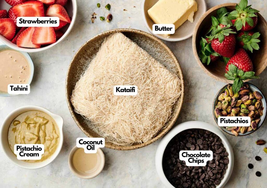 A bowl of kataifi surrounded by labeled ingredients: strawberries, butter, pistachios, chocolate chips, coconut oil, pistachio cream, and tahini for a delicious Dubai Chocolate Strawberry Cup.