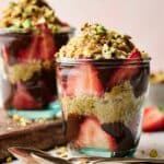 Two glass cups filled with layers of sliced strawberries, granola, chocolate, and chopped pistachios create a tempting Dubai Chocolate Strawberry Cup, with a spoon in front for easy indulging.