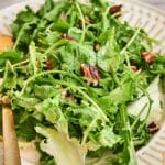 A plate of green salad with arugula, lettuce, and chopped pecans is served with a gold-colored fork.