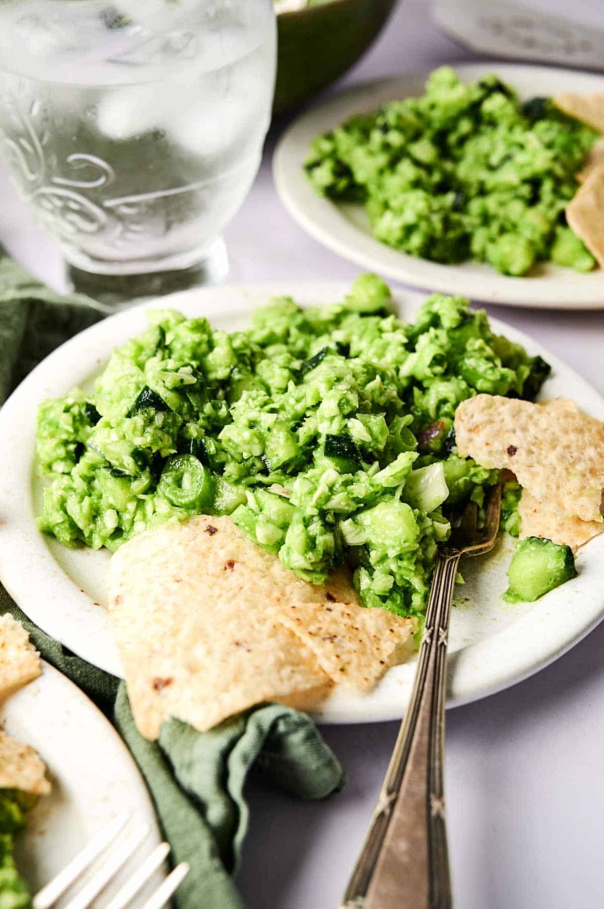 A plate of green goddess salad with tortilla chips and a fork, next to a glass of ice water and another fresh salad plate in the background.