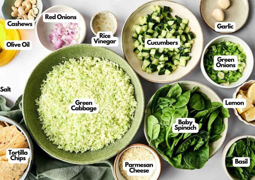 Various labeled ingredients for a green goddess salad, including chopped green cabbage, cucumbers, baby spinach, red and green onions, cashews, olive oil, and more, arranged in bowls on a white surface.