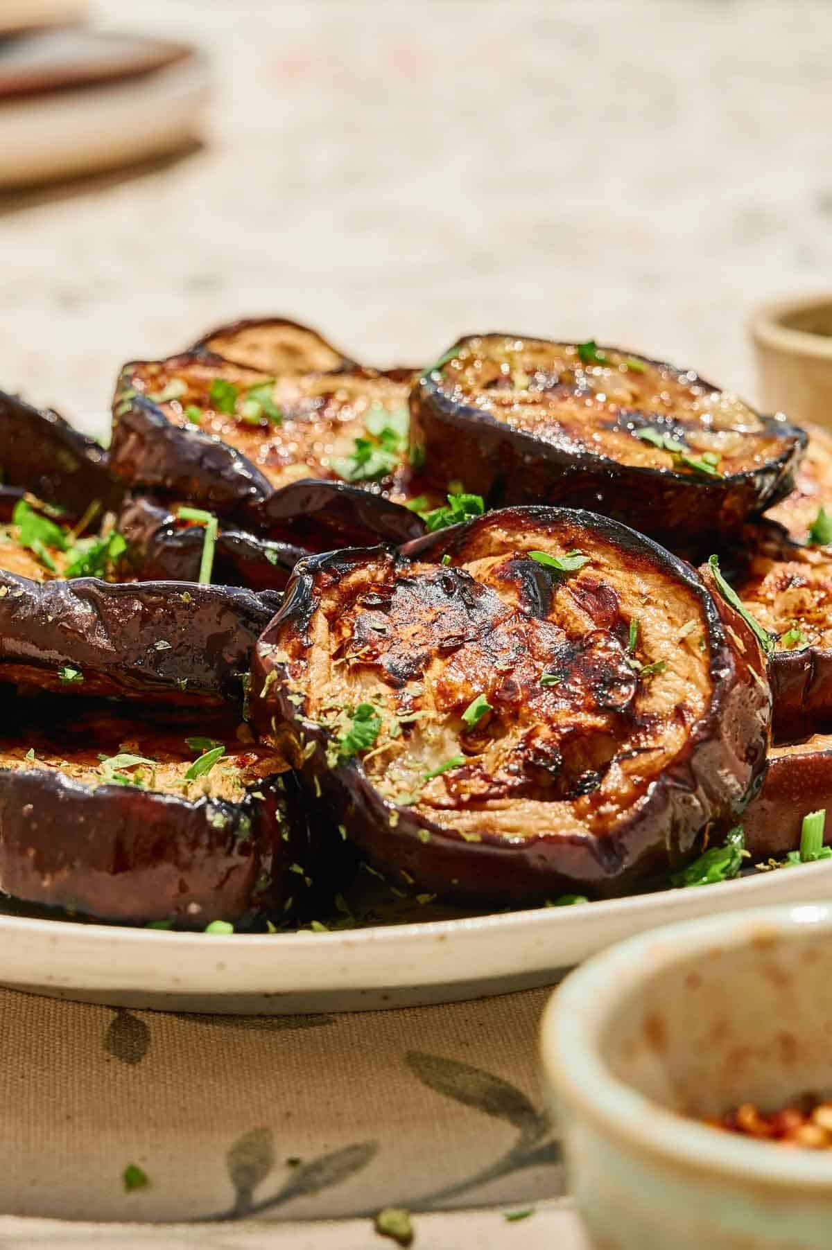 A plate of grilled eggplant slices, garnished with chopped herbs, is beautifully presented on a light-colored dish&mdash;a delightful way to enjoy the flavors of grilled eggplant.