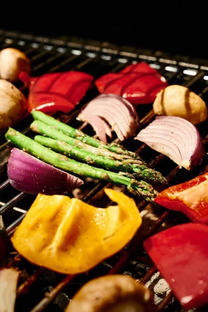 Grilled vegetables like asparagus, mushrooms, red onion, and bell peppers sizzle on a barbecue grate, creating a colorful and flavorful medley.