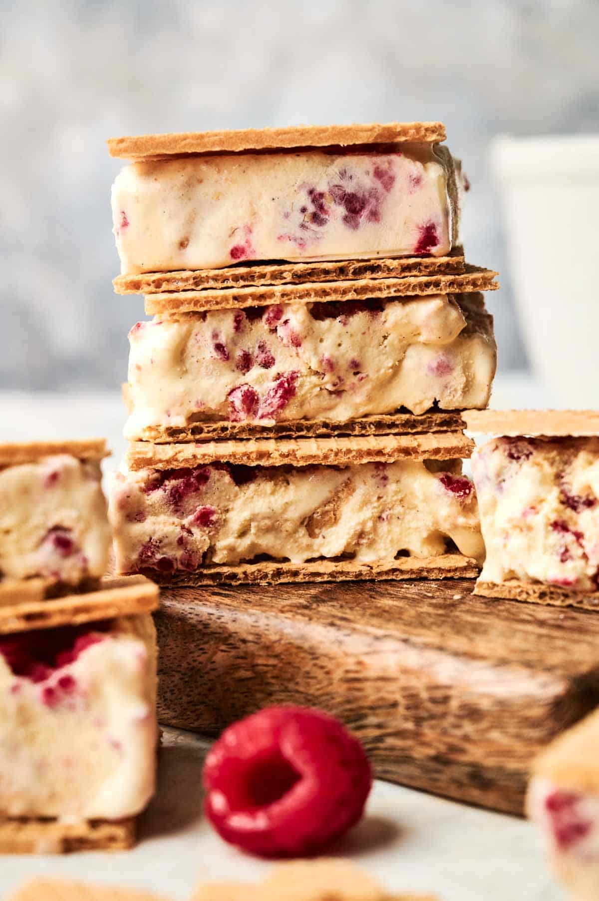 Ice cream sandwiches with raspberries are stacked on a wooden board, highlighting the irresistible layers of each Ice Cream Sandwich, with a single fresh raspberry in the foreground.