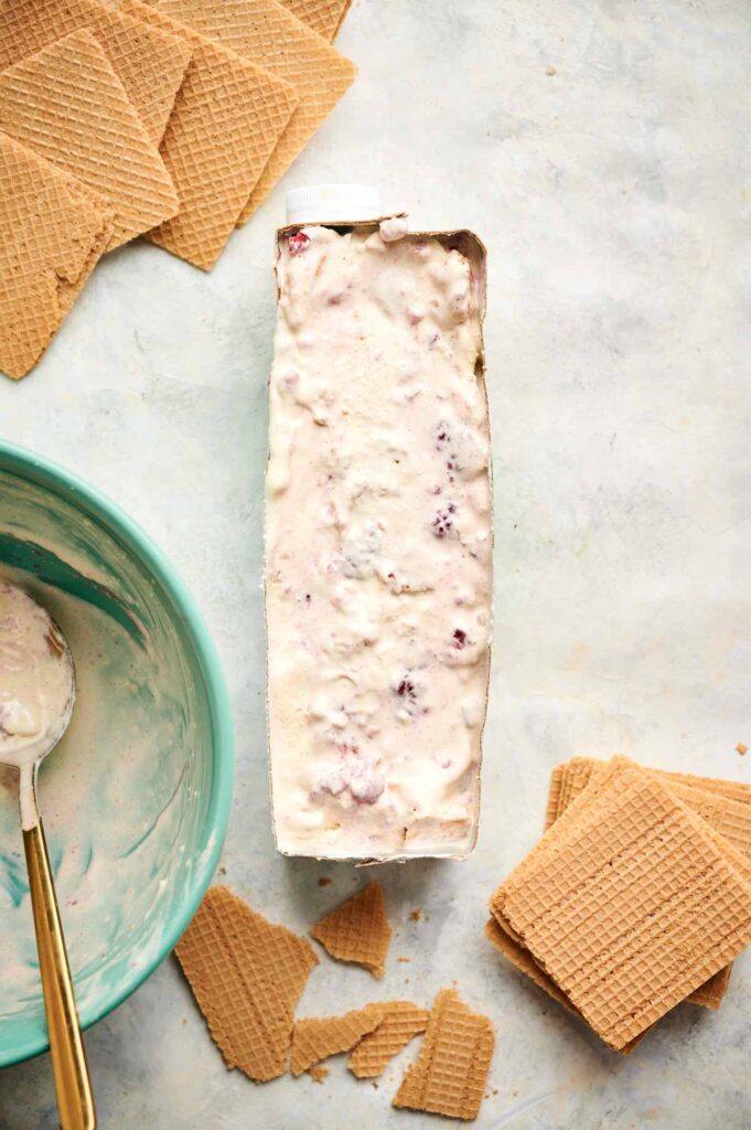 A rectangular container filled with a creamy ice cream sandwich mixture, possibly blended with berries, is surrounded by wafers and a large spoon in a turquoise bowl.