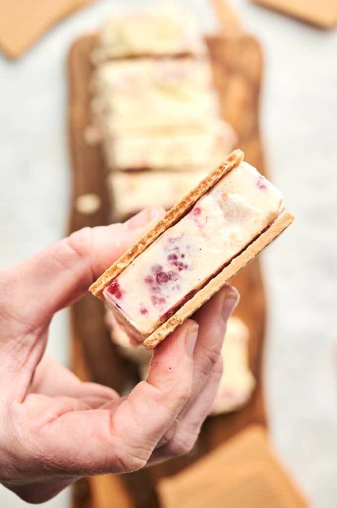 A hand holds a delicious Ice Cream Sandwich with visible berry pieces, made with graham crackers and creamy ice cream, while more tempting sandwiches rest on a wooden board in the background.