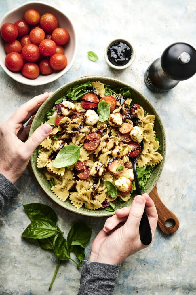 A person holds a bowl of Italian pasta salad with cherry tomatoes, mozzarella, basil, and balsamic glaze, fork in hand. A bowl of cherry tomatoes and a pepper grinder are nearby.