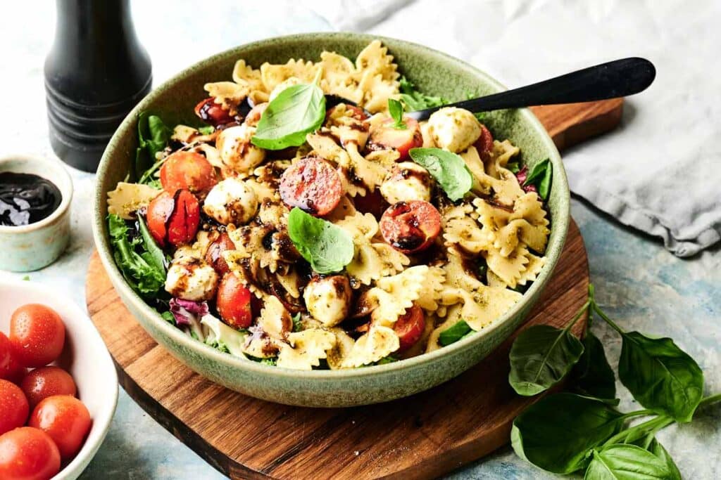 A bowl of Italian pasta salad with farfalle, cherry tomatoes, mozzarella balls, fresh basil, and balsamic drizzle sits on a wooden board, surrounded by spinach leaves and a bowl of tomatoes.