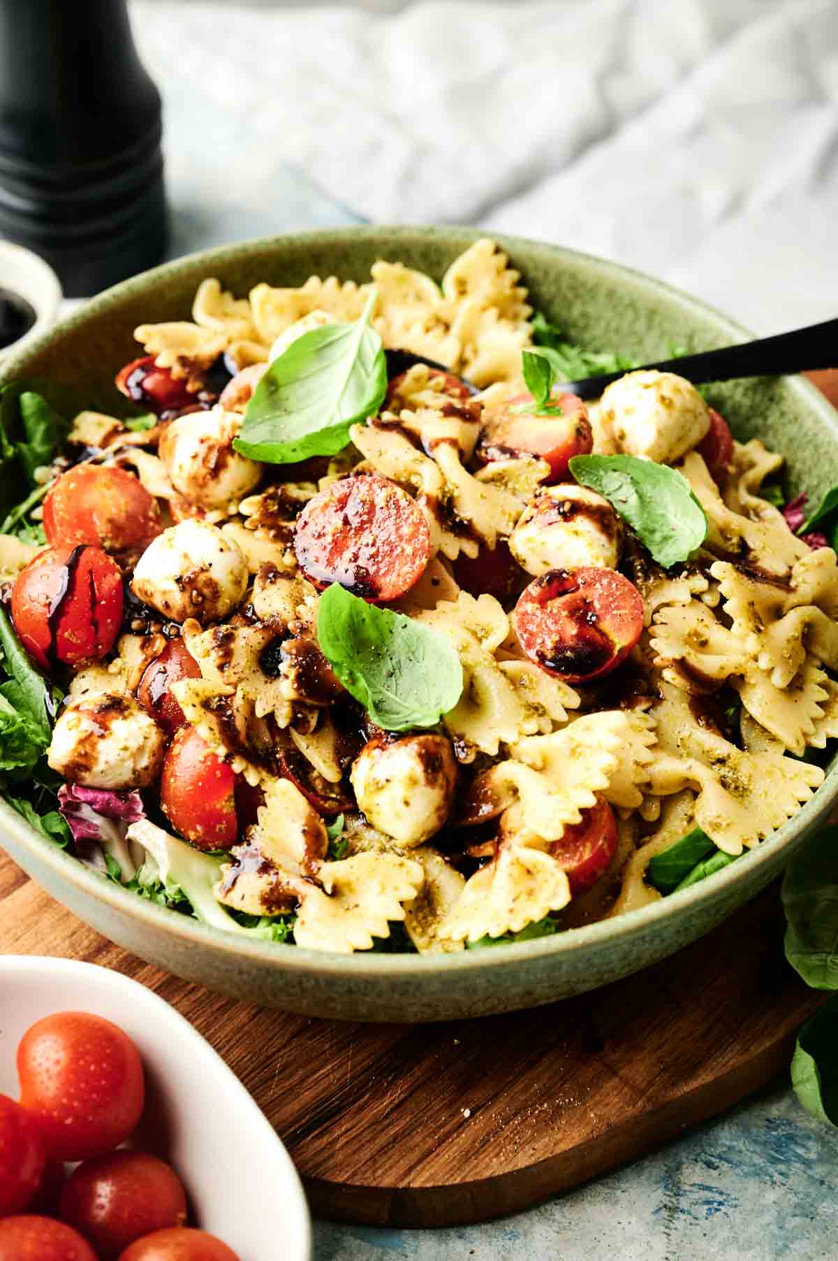 A bowl of Italian pasta salad featuring bowtie pasta, cherry tomatoes, mozzarella balls, fresh basil leaves, and a drizzle of balsamic dressing on top.