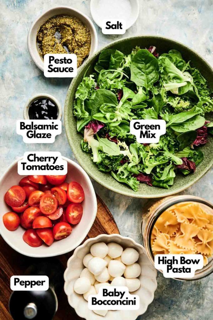 A bowl of Italian pasta salad mix surrounded by labeled ingredients: pesto sauce, salt, balsamic glaze, cherry tomatoes, pepper, baby bocconcini, and bow tie pasta.