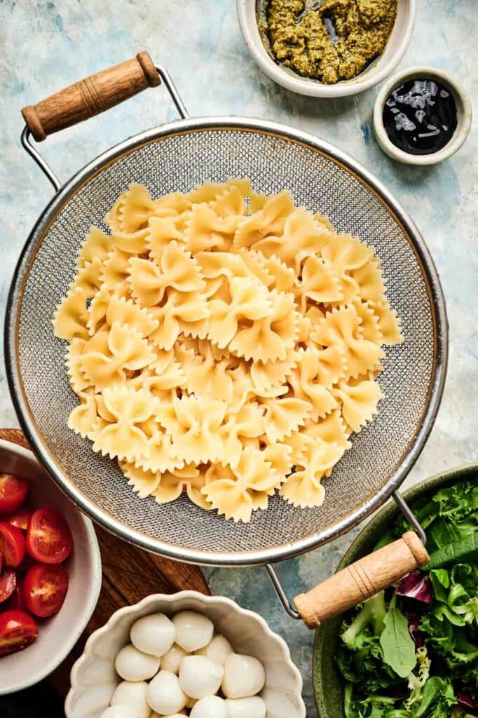 A colander filled with cooked farfalle pasta is surrounded by bowls of pesto, olives, cherry tomatoes, mozzarella balls, and mixed greens&mdash;everything you need to create a fresh Italian pasta salad.