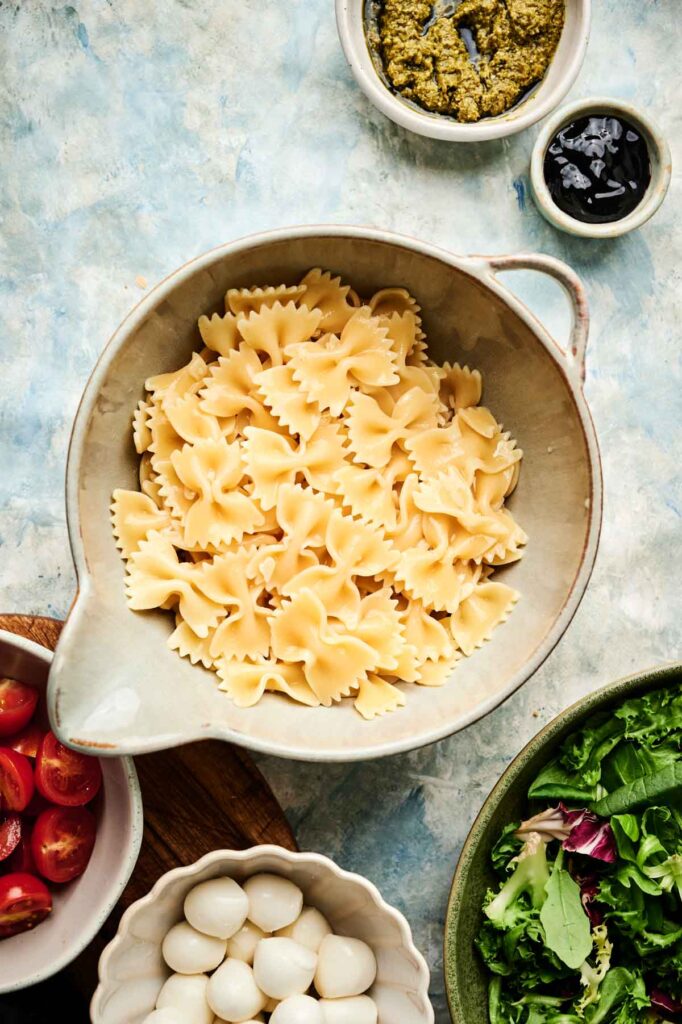 A bowl of cooked farfalle pasta is surrounded by small bowls of pesto, black olives, cherry tomatoes, mozzarella balls, and mixed salad greens&mdash;perfect ingredients for an Italian pasta salad.