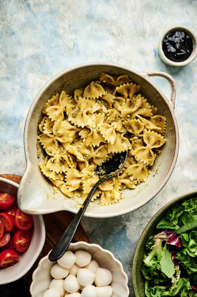 A bowl of farfalle pasta mixed with pesto creates a vibrant Italian pasta salad, surrounded by bowls of cherry tomatoes, mozzarella balls, mixed greens, and a small dish of balsamic vinegar.
