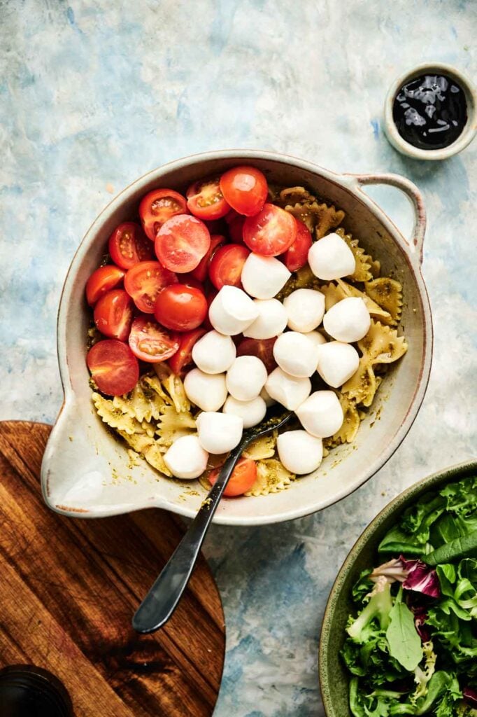 A bowl of Italian pasta salad with bowtie pasta, halved cherry tomatoes, and mozzarella balls, a metal spoon inside; salad and a small bowl of dressing are nearby.