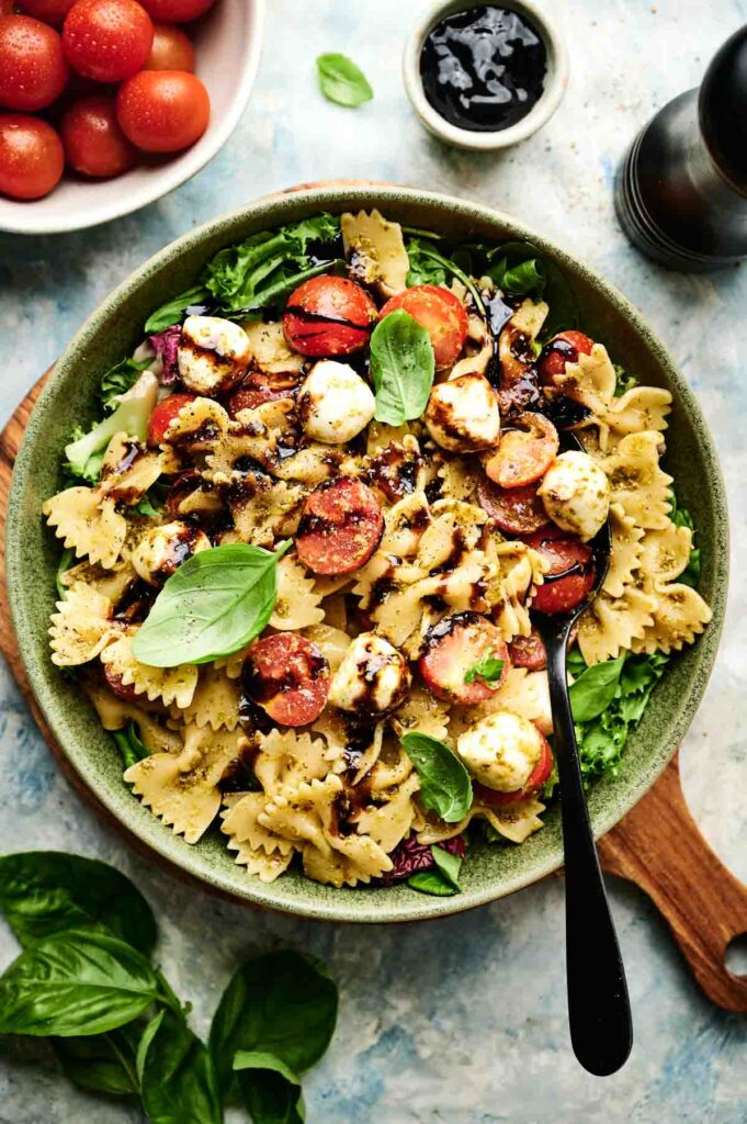 A bowl of Italian pasta salad featuring bowtie pasta, cherry tomatoes, mozzarella balls, fresh basil, and balsamic glaze, served with a fork and garnished with leafy greens.