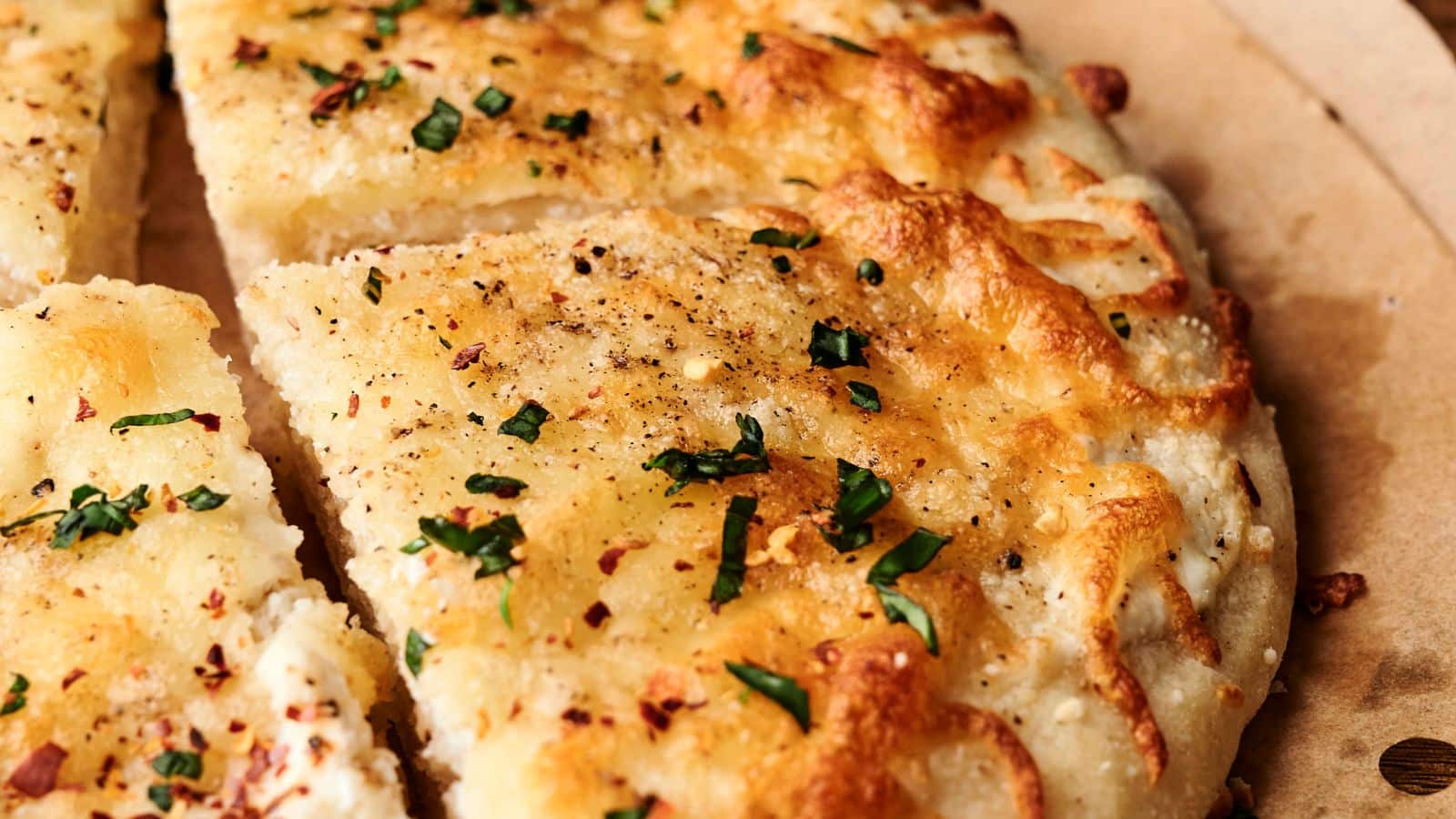 Close-up of a sliced, golden-brown focaccia bread topped with herbs, black pepper, and red pepper flakes on parchment paper.