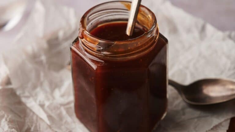 A hexagonal glass jar filled with dark brown sauce, with a metal spoon in the jar and another spoon resting nearby on crumpled parchment paper.
