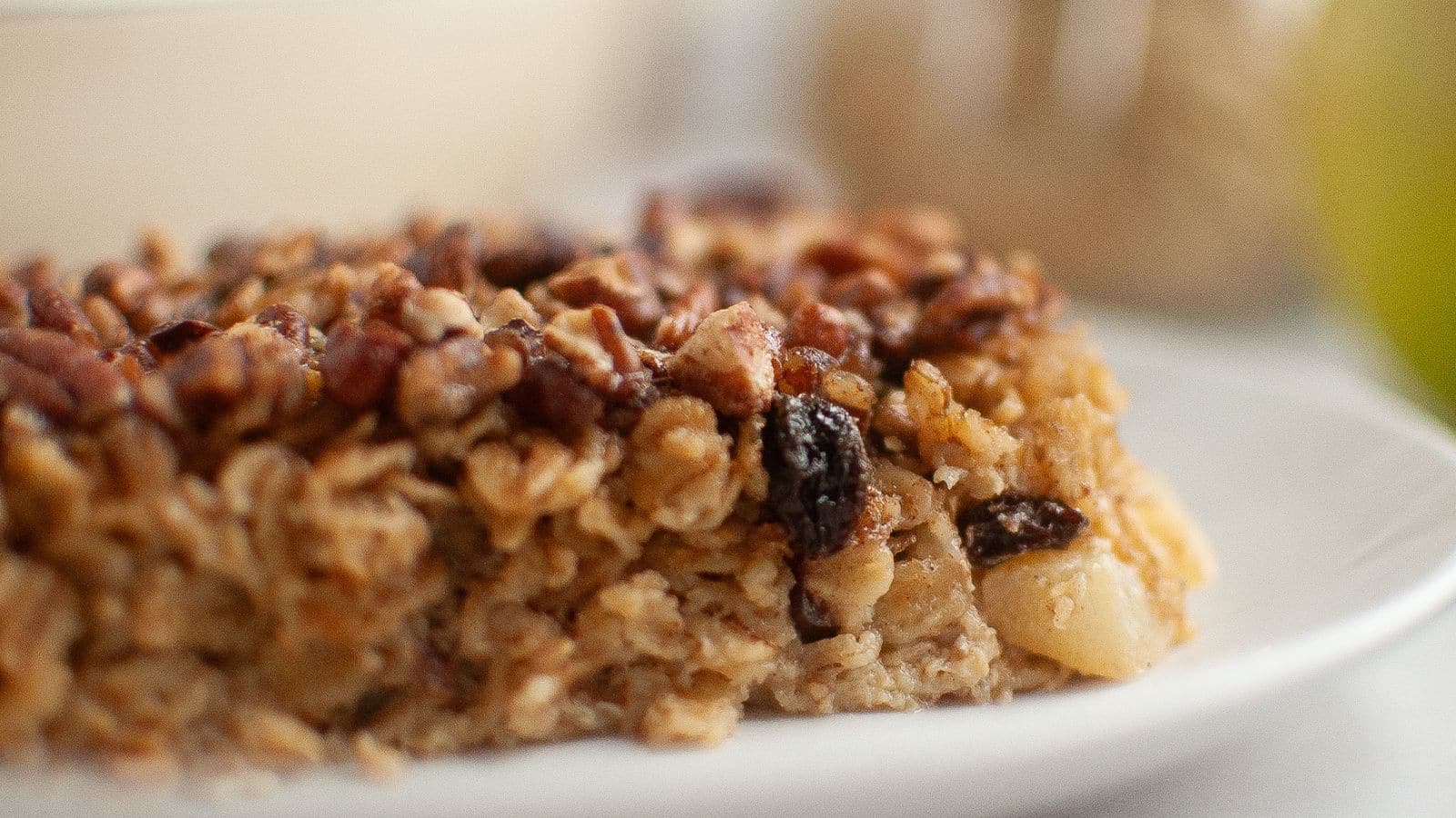A close-up of a baked oatmeal bar with visible oats, nuts, and raisins on a white plate.