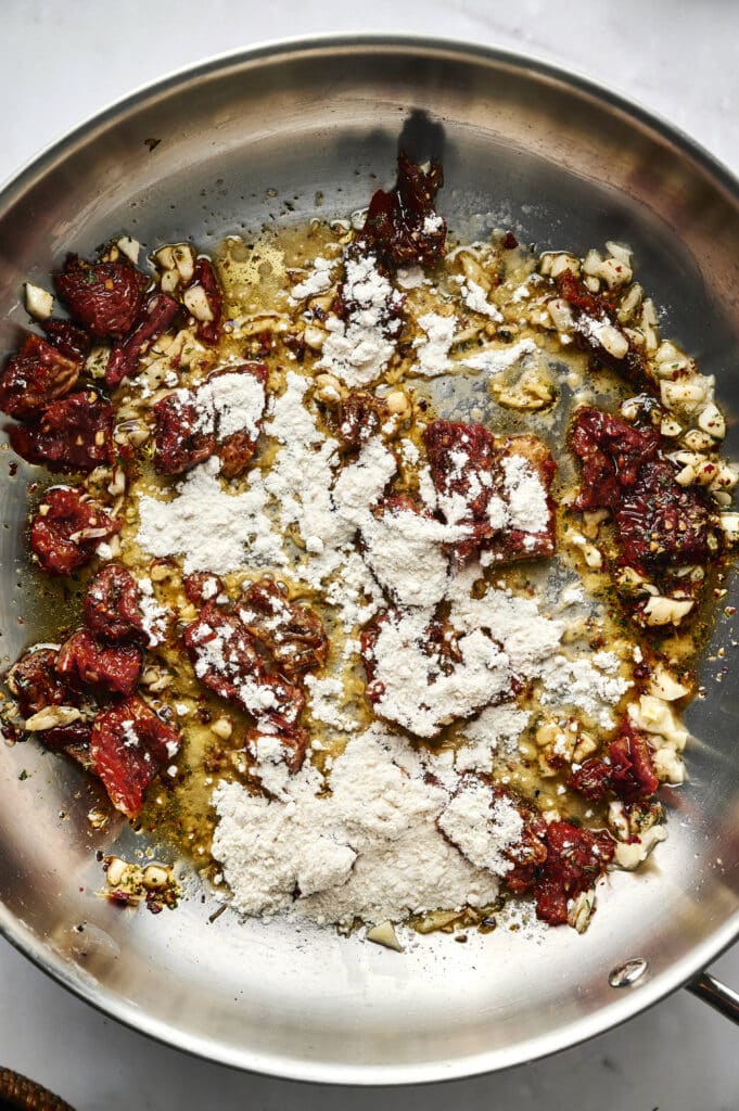 A stainless steel pan containing chopped sun-dried tomatoes, minced garlic, spices, oil, and a layer of flour sprinkled on top is being sautéed for a flavorful Marry Me Tortellini dish.