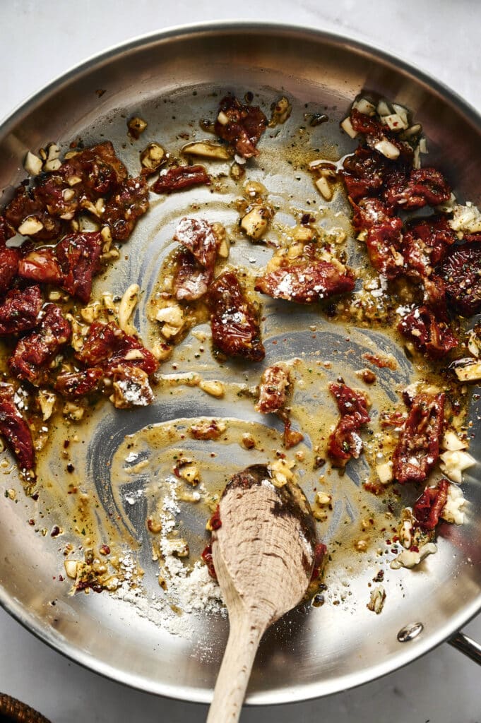 A stainless steel pan with sautéed sun-dried tomatoes, chopped garlic, and oil for Marry Me Tortellini, stirred gently with a wooden spoon.