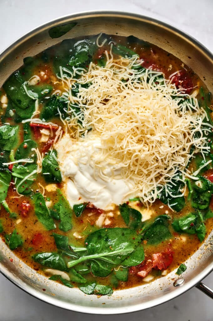 A skillet containing Marry Me Tortellini soup with spinach, tomatoes, a dollop of sour cream, and a pile of shredded cheese on top, before mixing.