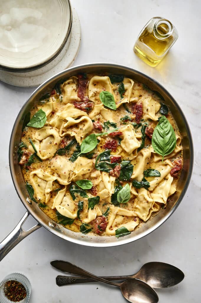 A skillet filled with creamy Marry Me Tortellini pasta, sun-dried tomatoes, spinach, and basil leaves sits on a marble surface beside a bottle of olive oil and a stack of plates.