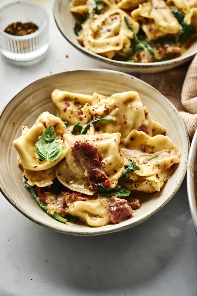 A bowl of creamy Marry Me Tortellini pasta with sun-dried tomatoes, spinach, and visible herbs, served on a white surface.
