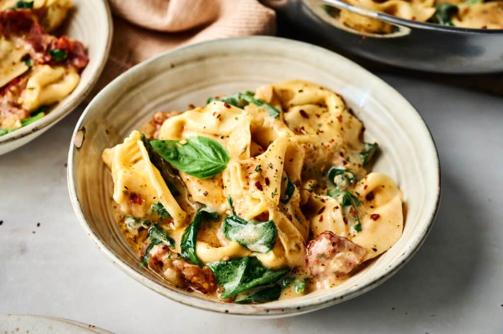 A bowl of creamy Marry Me Tortellini pasta with spinach, sun-dried tomatoes, and a basil garnish.