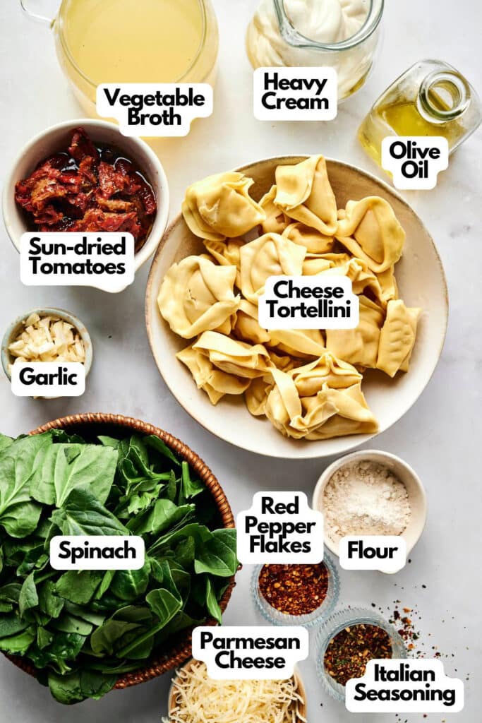 A flat lay of ingredients for Marry Me Tortellini, including cheese tortellini, spinach, garlic, sun-dried tomatoes, vegetable broth, heavy cream, olive oil, flour, red pepper flakes, parmesan cheese, and Italian seasoning.