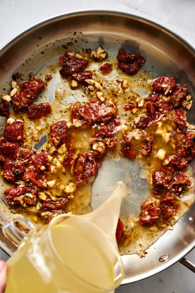 A metal skillet with sautéed sun-dried tomatoes and garlic, as broth is poured in from a clear pitcher—perfect for crafting Marry Me Tortellini.