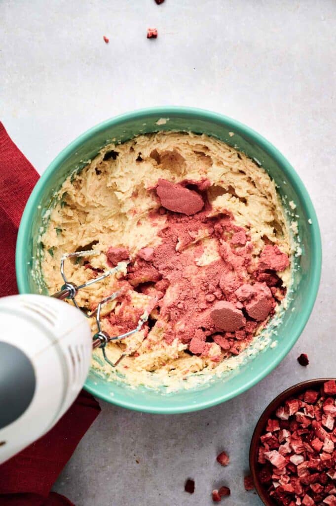 A hand mixer blending strawberry cookies dough in a green bowl with pink powder and chopped red pieces on top, plus a small bowl of chopped red pieces beside it.