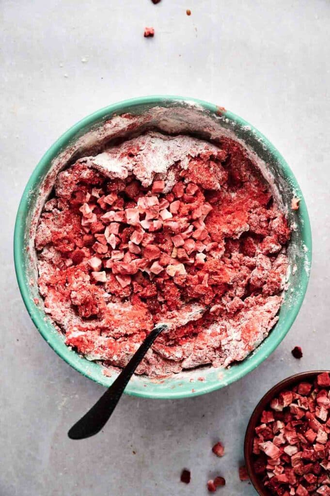 A green mixing bowl containing flour and chopped strawberries for strawberry cookies, with a black spoon inside. A small bowl with more chopped strawberries sits beside it.