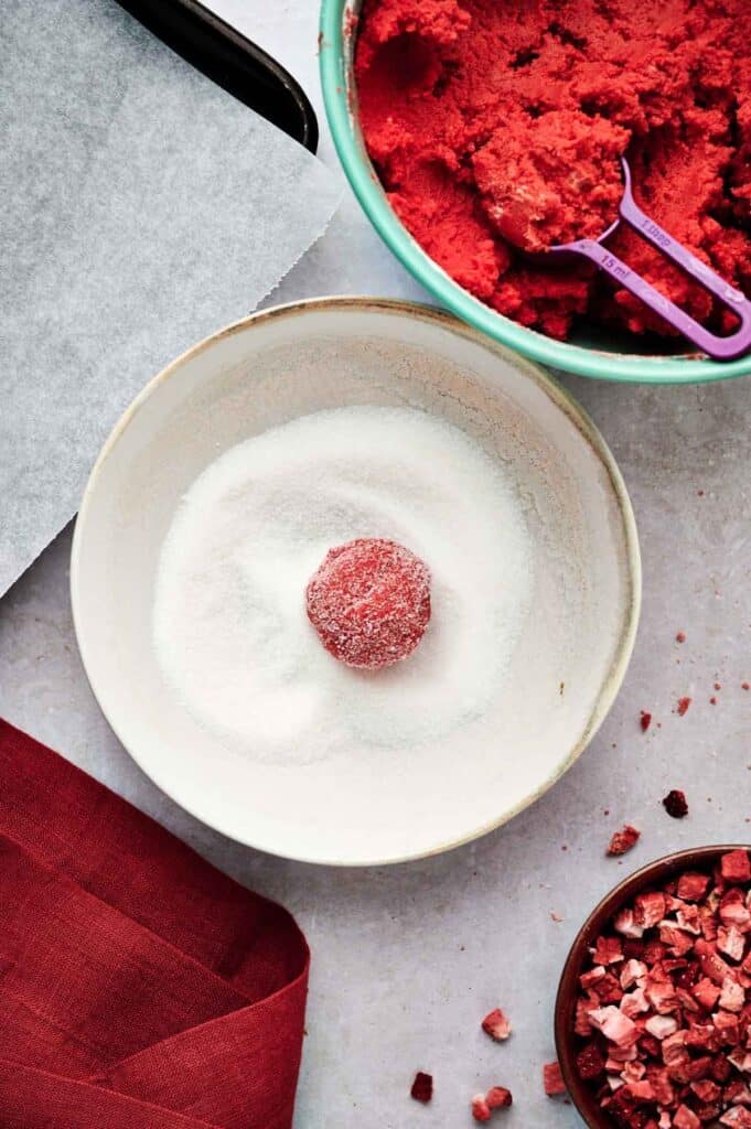 A red dough ball for strawberry cookies is being rolled in a bowl of white sugar, with extra red dough and mix-ins nearby on the countertop.