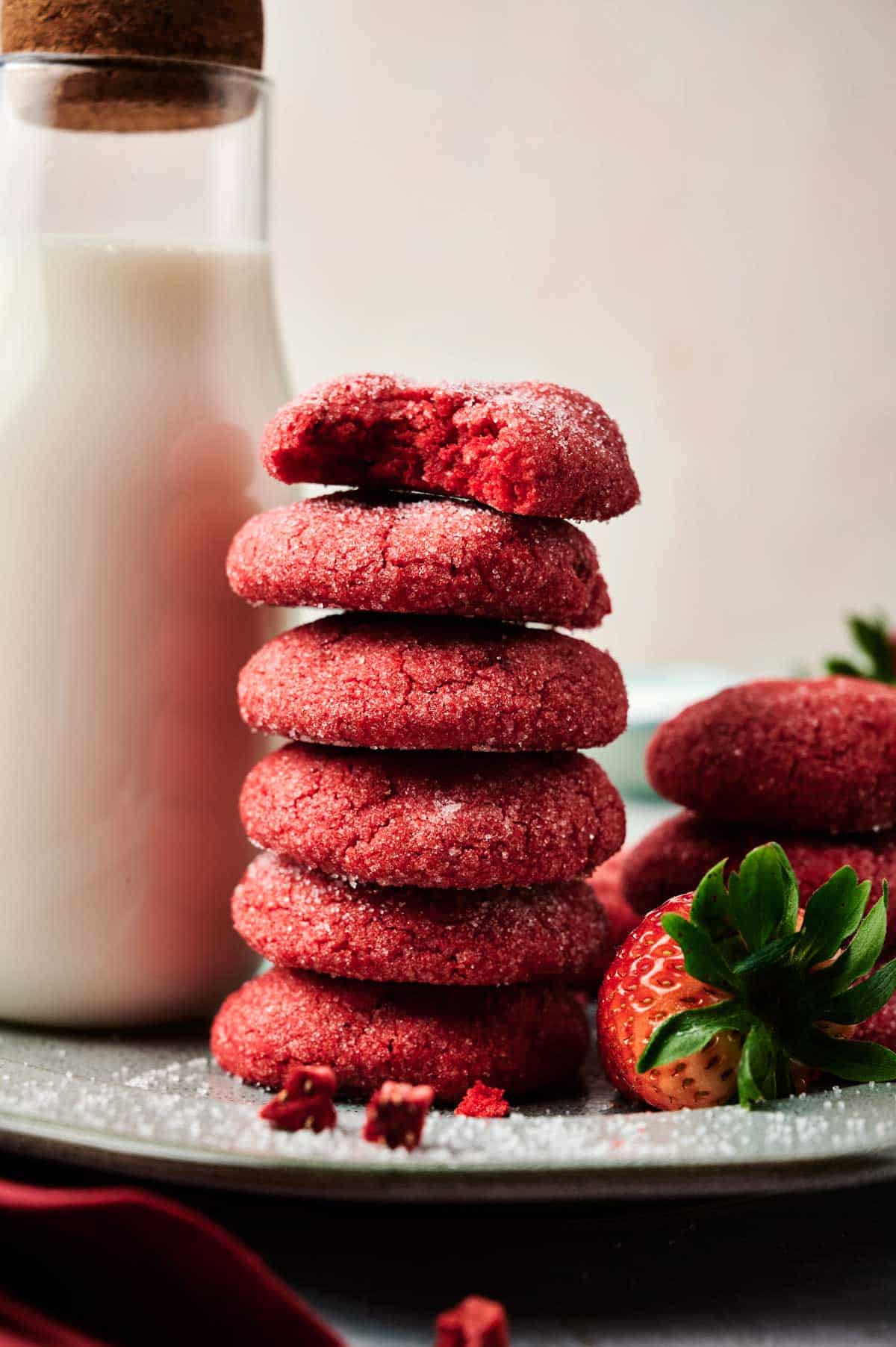 A stack of red sugar-coated strawberry cookies, one with a bite taken out, sits beside a glass bottle of milk and fresh strawberries on a plate.