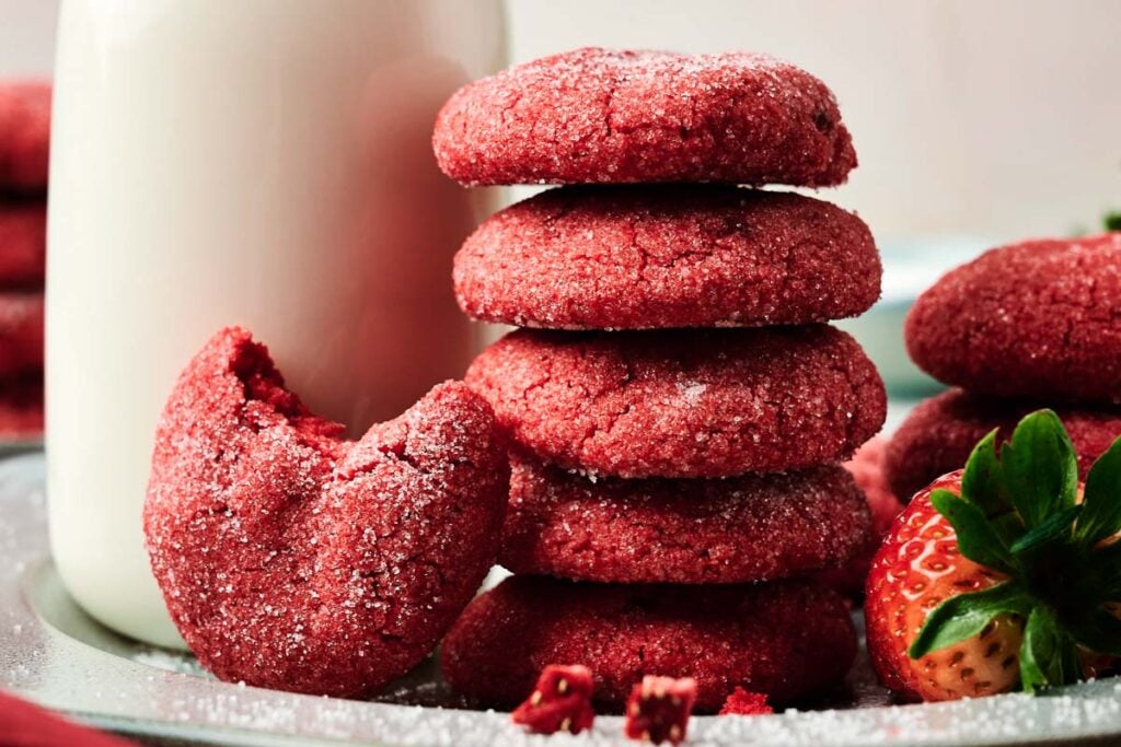 A stack of red sugar-coated strawberry cookies sits next to a bitten cookie, a fresh strawberry, and a glass of milk on a plate.