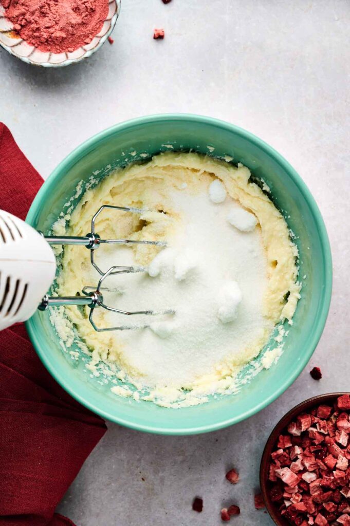An electric hand mixer blends sugar and butter in a green bowl, with bowls of red powder for strawberry cookies and chopped ingredients nearby on a light surface.