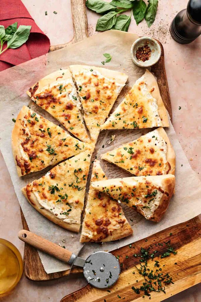 A sliced White Pizza on parchment paper, garnished with chopped herbs. A pizza cutter and knife rest nearby, with fresh basil leaves and seasonings in the background.