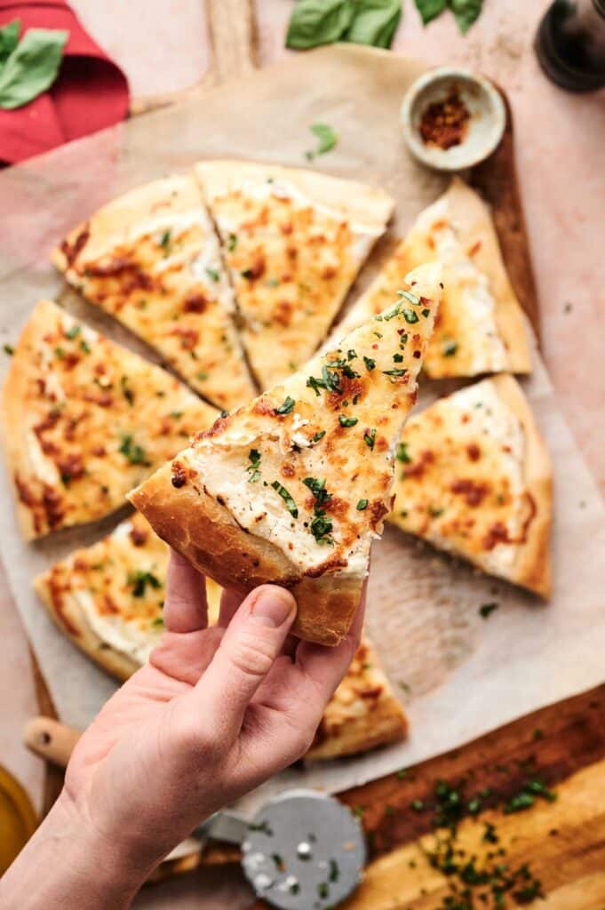A hand holds a slice of cheesy White Pizza topped with fresh herbs, with the rest of the delicious pizza sliced on a wooden board in the background.