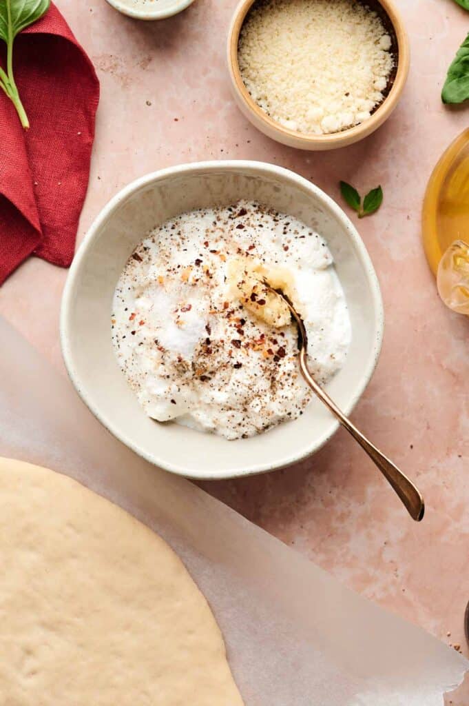 A bowl of ricotta cheese mixed with seasonings and a spoon, surrounded by grated cheese, olive oil, herbs, a red napkin, and pizza dough—perfect for crafting a delicious white pizza—on a pink surface.